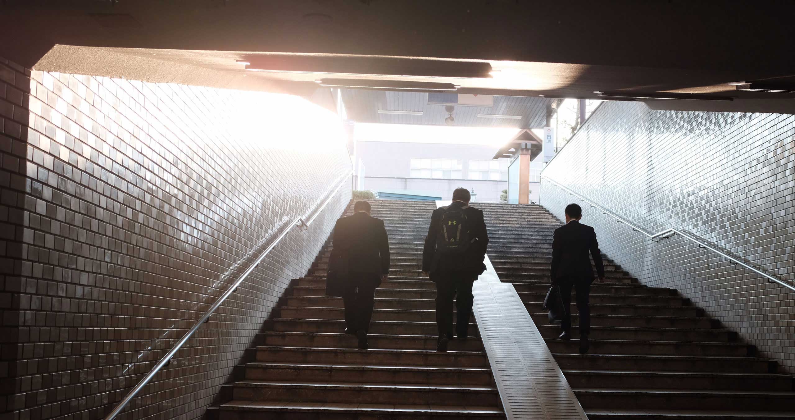 Three businessmen ascend the subway stairs towards daylight
